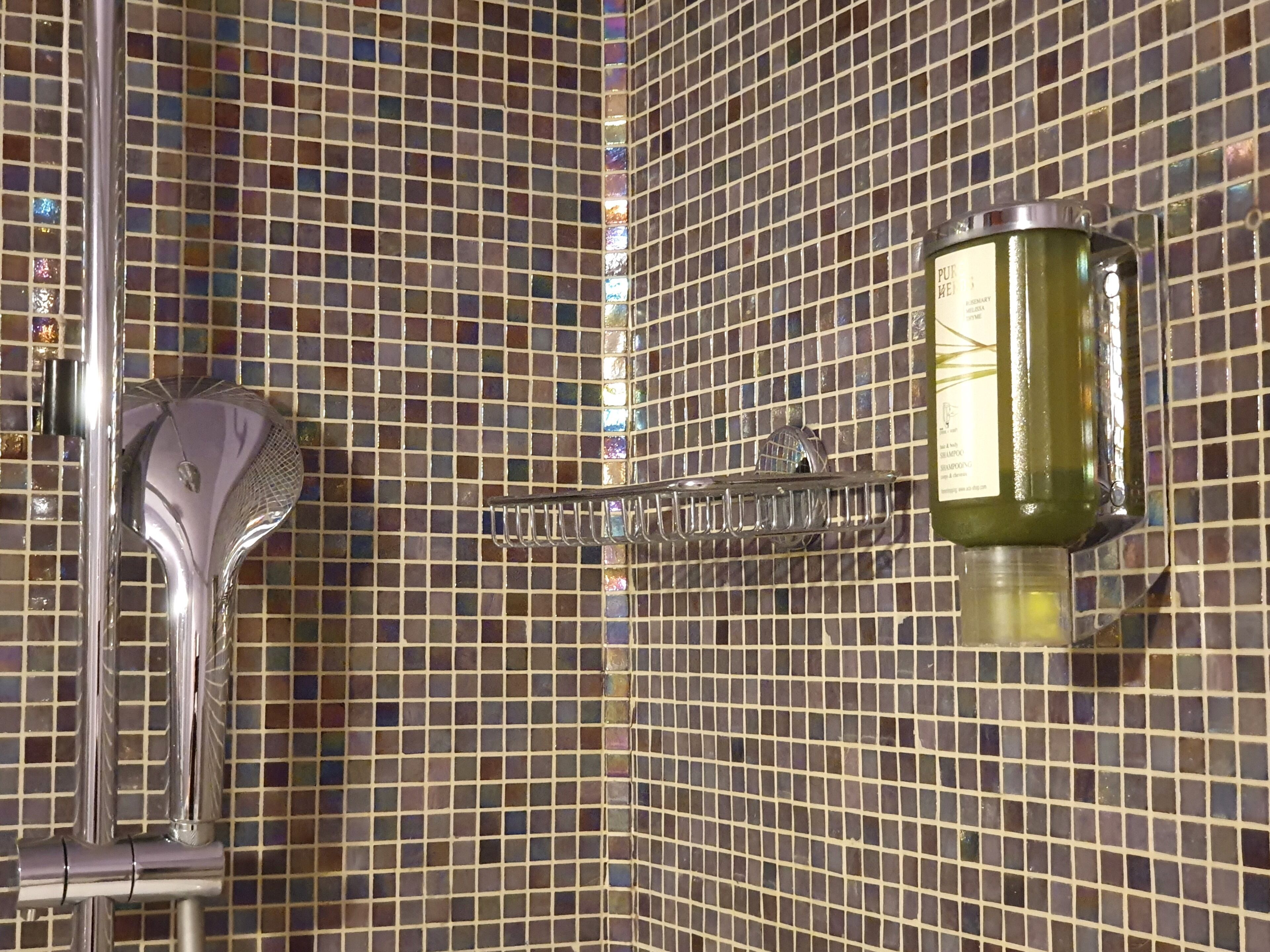 Une salle de bain avec un mur recouvert d’une mosaïque, un robinet chromé et une bouteille de shampoing posée sur un porte-savon fixé au mur.