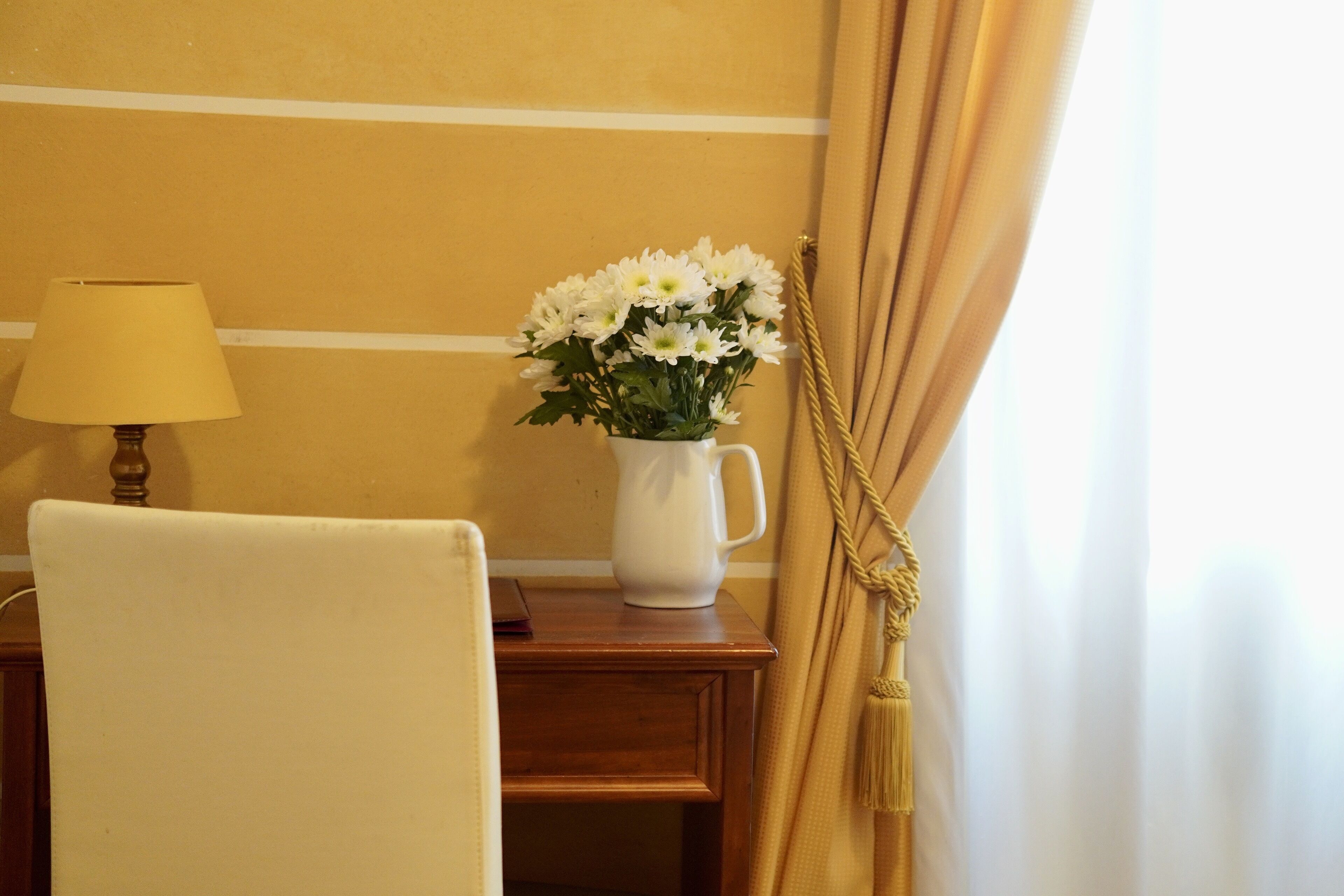 Una habitación con una mesita de noche de madera, una lámpara blanca y un jarrón con flores blancas.