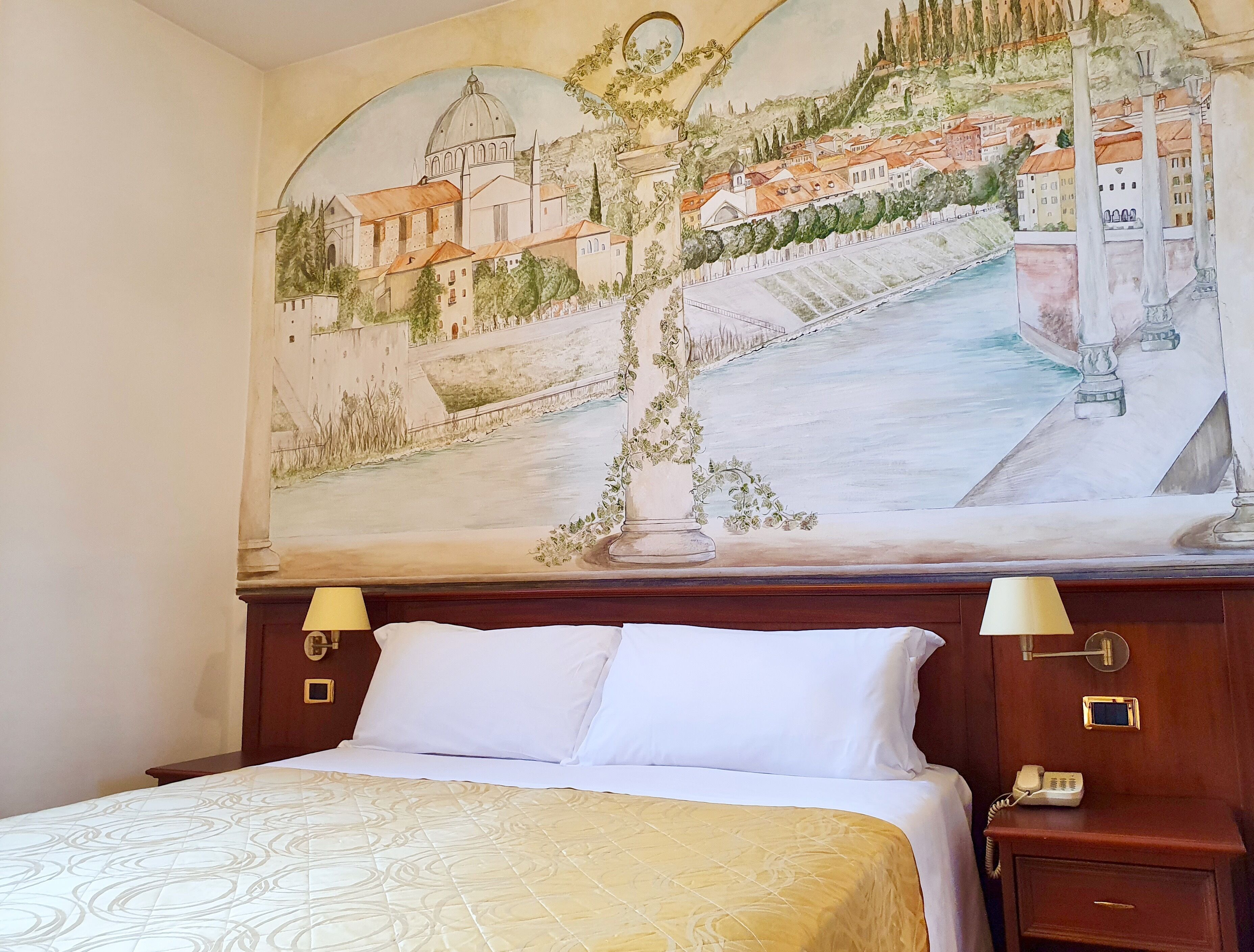 A hotel room with a large mural of a cityscape on the wall, a bed with white linens, and a bedside table with a lamp and a phone.