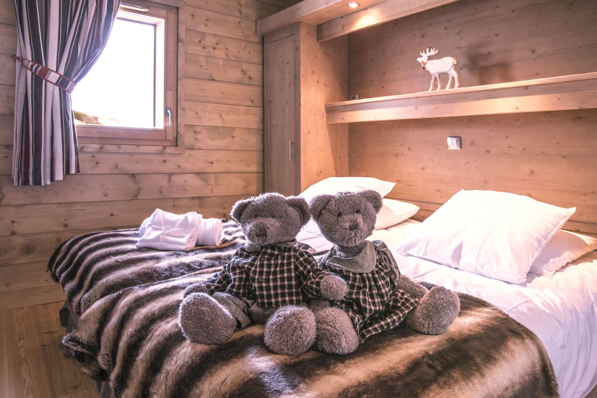 Une pièce de cabane en bois comprenant un lit, deux ours en peluche, une fenêtre avec des rideaux et une étagère sur laquelle est posée une figurine d’élan.