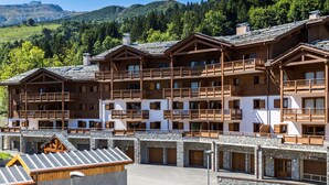 A multi-story wooden building with balconies and a stone facade, set against a mountainous backdrop. - CGH  La Grange aux Fées (Les Avanchers-Valmorel)