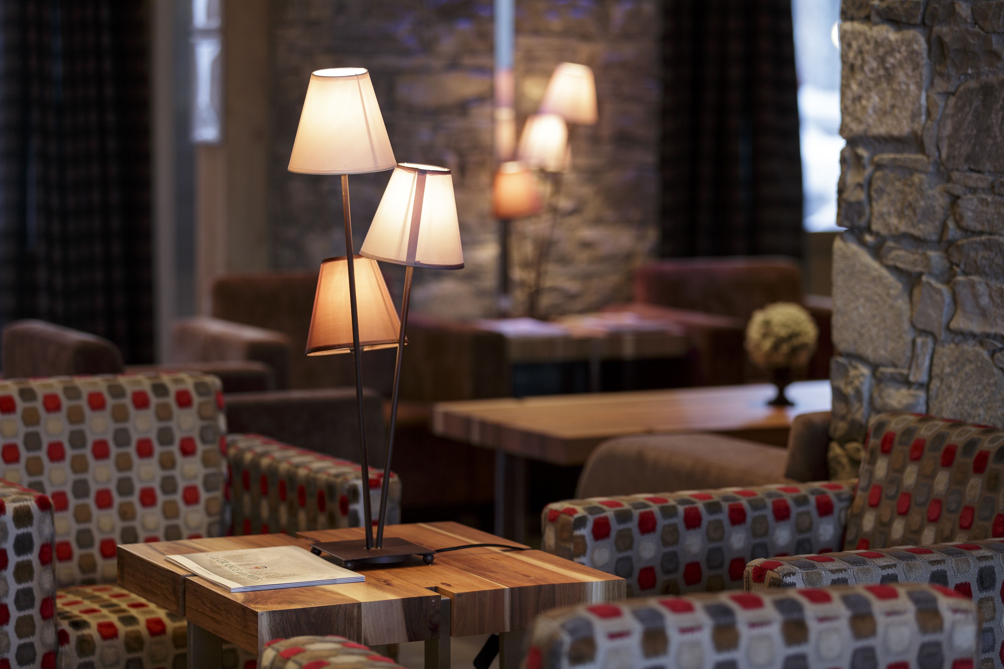 A cozy seating area with patterned upholstery, a wooden table with a lamp, and a stone wall in the background.