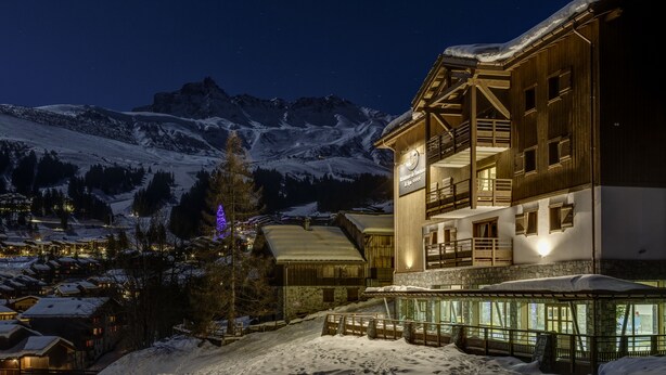 A snow-covered mountain town at night with a prominent wooden building featuring balconies and a lit sign. - CGH  La Grange aux Fées (Les Avanchers-Valmorel)