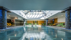 A large indoor swimming pool with a skylight, stone columns, and green lounge chairs. - CGH Résidences Boutique Le Napoléon (Montgenevre)