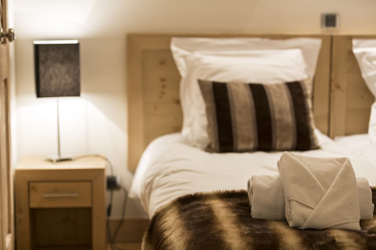 A neatly made bed with white and striped pillows, a bedside lamp, and a folded towel on a brown furry blanket.