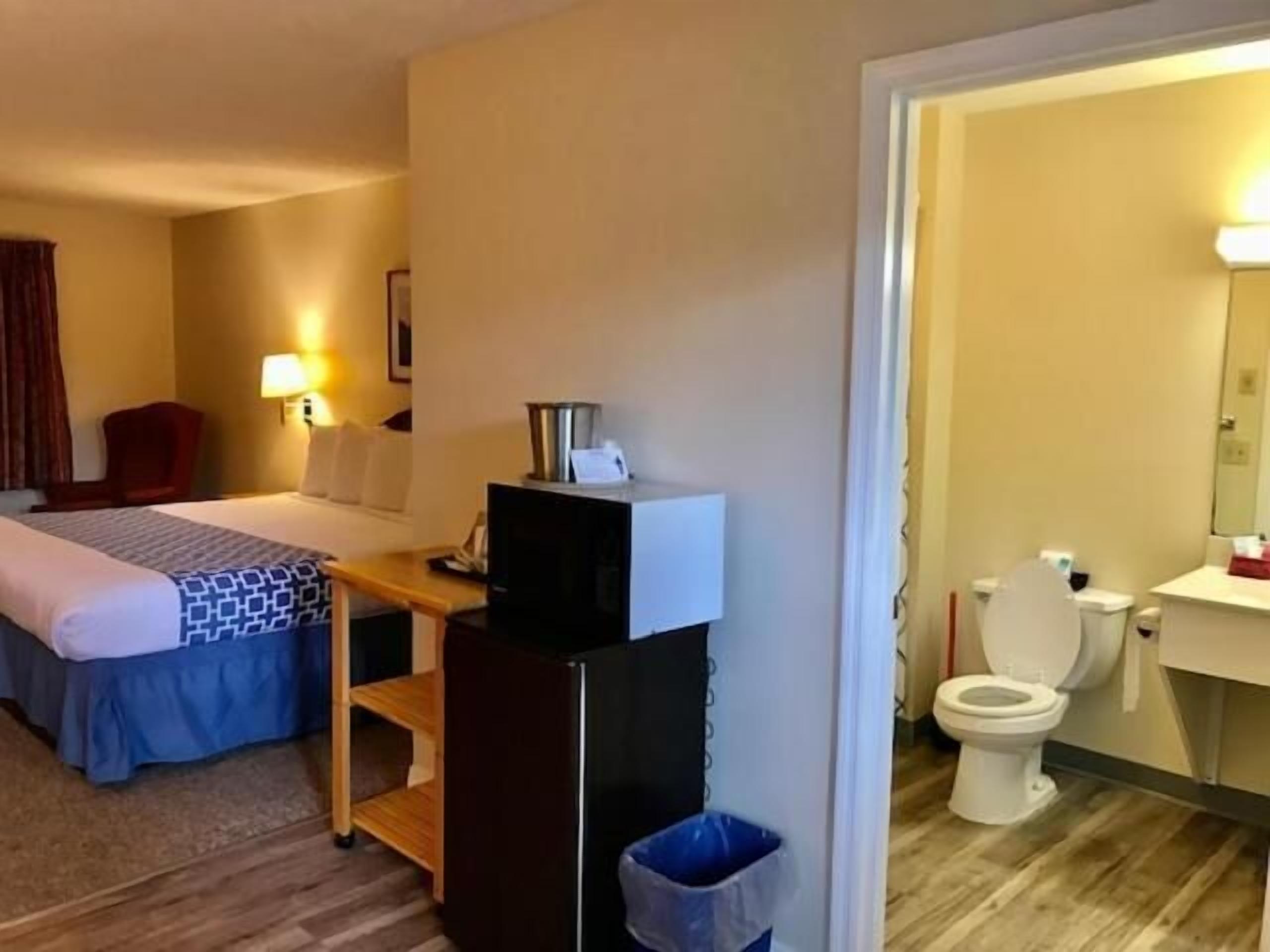 A hotel room with a bed, a desk with a computer, a trash bin, and a bathroom with a toilet and sink.