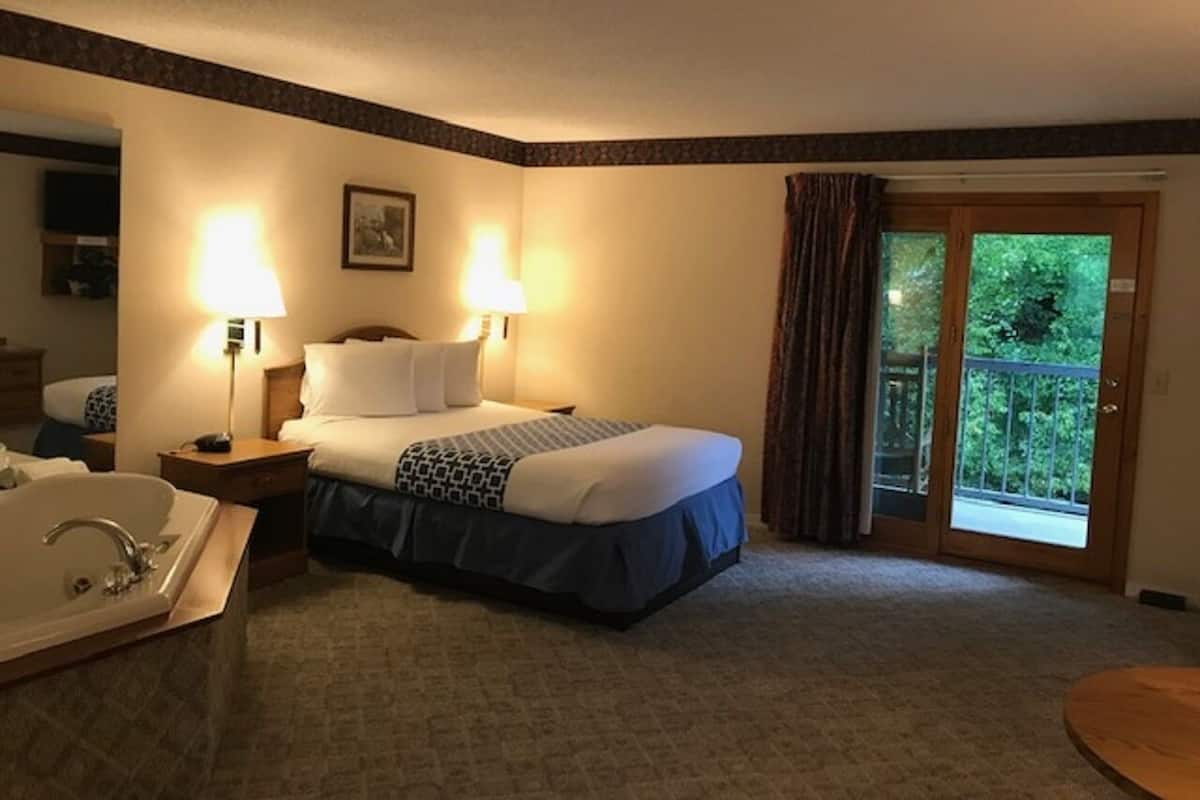 A hotel room with a bed, two bedside lamps, a bathroom with a sink and mirror, and a view of greenery through a sliding glass door.
