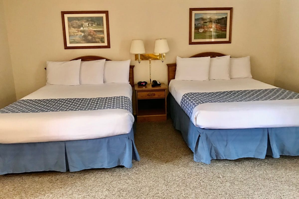 A hotel room with two beds, each with a blue bedspread and white linens. There are two framed pictures on the walls, a nightstand with a lamp and phone, and a carpeted floor.