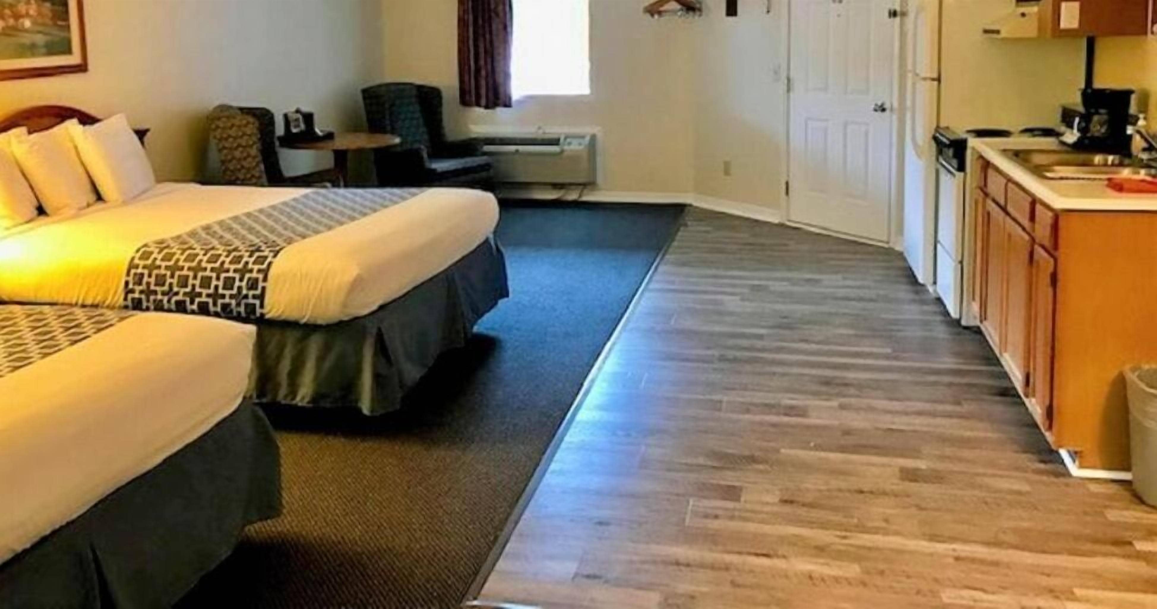 A hotel room with two beds, a small desk, a chair, a kitchenette, and a window with curtains.