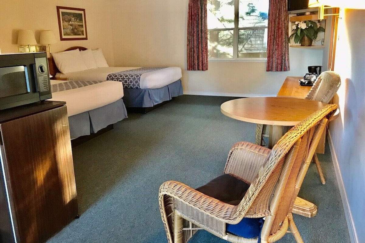 A compact hotel room with a bed, a small table, wicker chairs, a television, and a refrigerator.