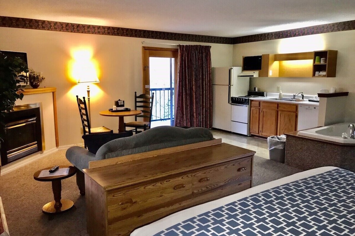 A hotel room with a bed, a sofa, a desk, a chair, a lamp, a kitchen area, and a bathroom.