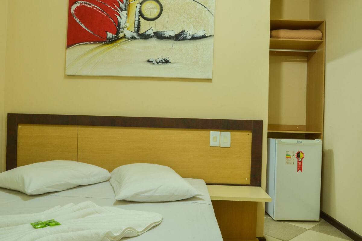 A hotel room with a bed, white pillows, a wall-mounted artwork, a small refrigerator, and a wooden shelf.