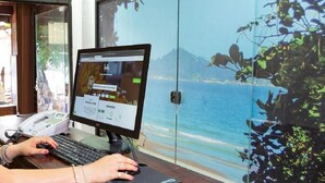 Monitor de computador exibindo um site, um telefone e vista para uma praia através de uma porta de vidro.