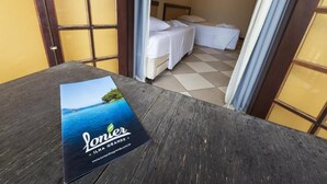 Uma mesa de madeira com um folheto intitulado "Lonier Ilha Grande" em um quarto com piso xadrez, duas camas e uma janela com cortinas.