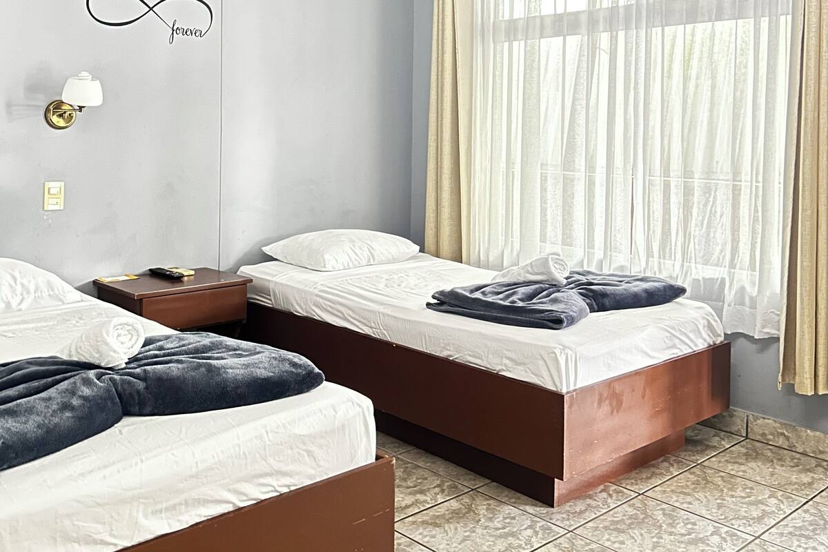 A bedroom with two beds, a nightstand, a wall-mounted lamp, and a window with sheer curtains.