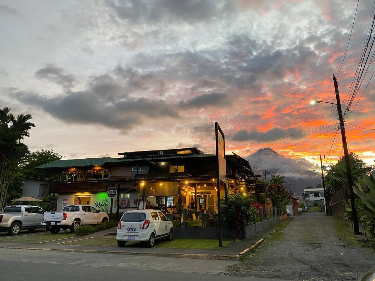A restaurant with outdoor seating, a mountain in the background, and parked cars in front.