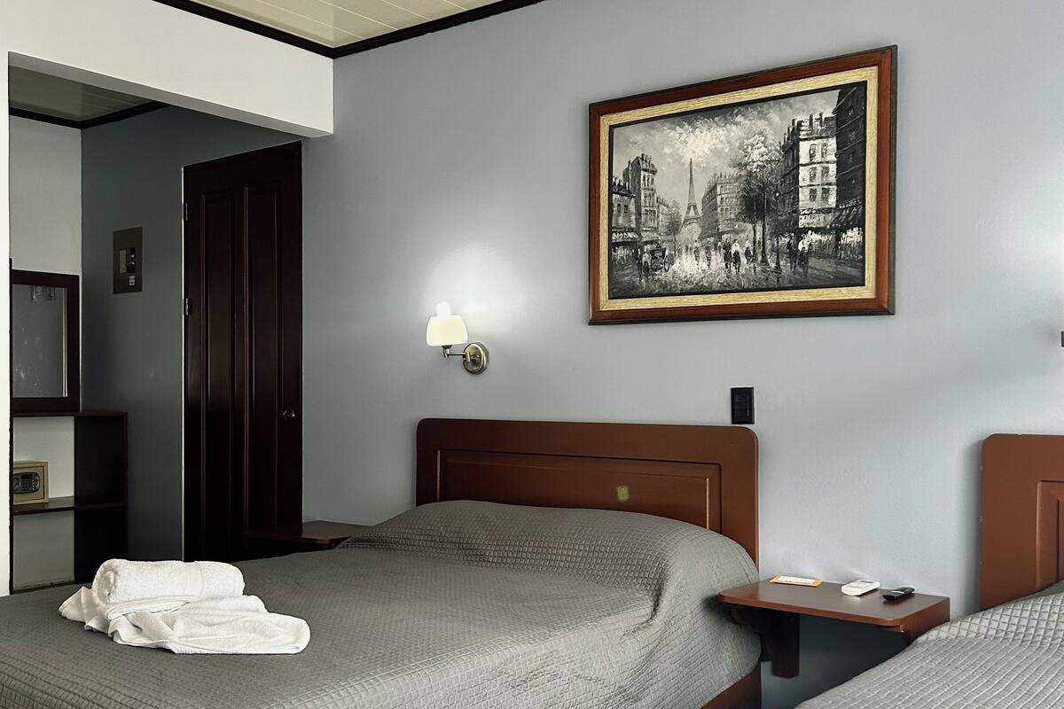 A hotel room with two beds, a painting on the wall, a bedside table with a lamp, and a door leading to another room.