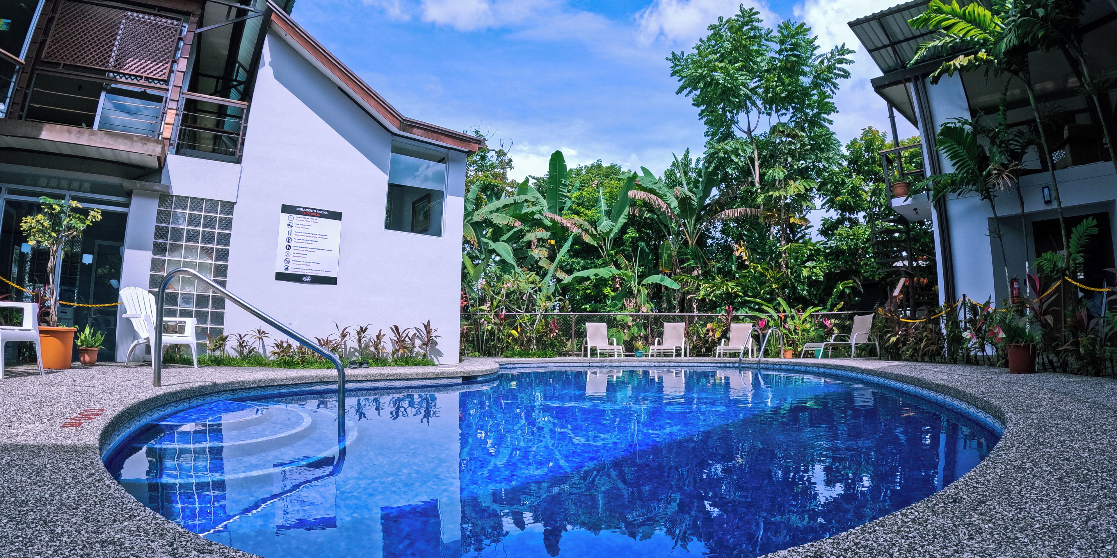 A swimming pool surrounded by lush greenery, with a building featuring a balcony and outdoor seating area.