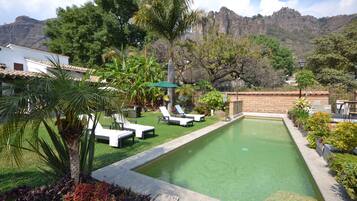 A swimming pool with lounge chairs, surrounded by greenery and a mountain backdrop.