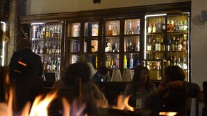 Un coin bar avec un feu de cheminée en premier plan, des étagères remplies de bouteilles et une personne debout au fond.