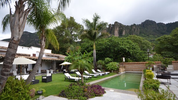 Un espace piscine doté de chaises longues et de parasols, entouré d’une végétation luxuriante et avec une chaîne de montagnes en toile de fond.