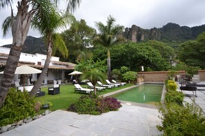 A pool area with lounge chairs and umbrellas, surrounded by lush greenery and a mountainous backdrop.