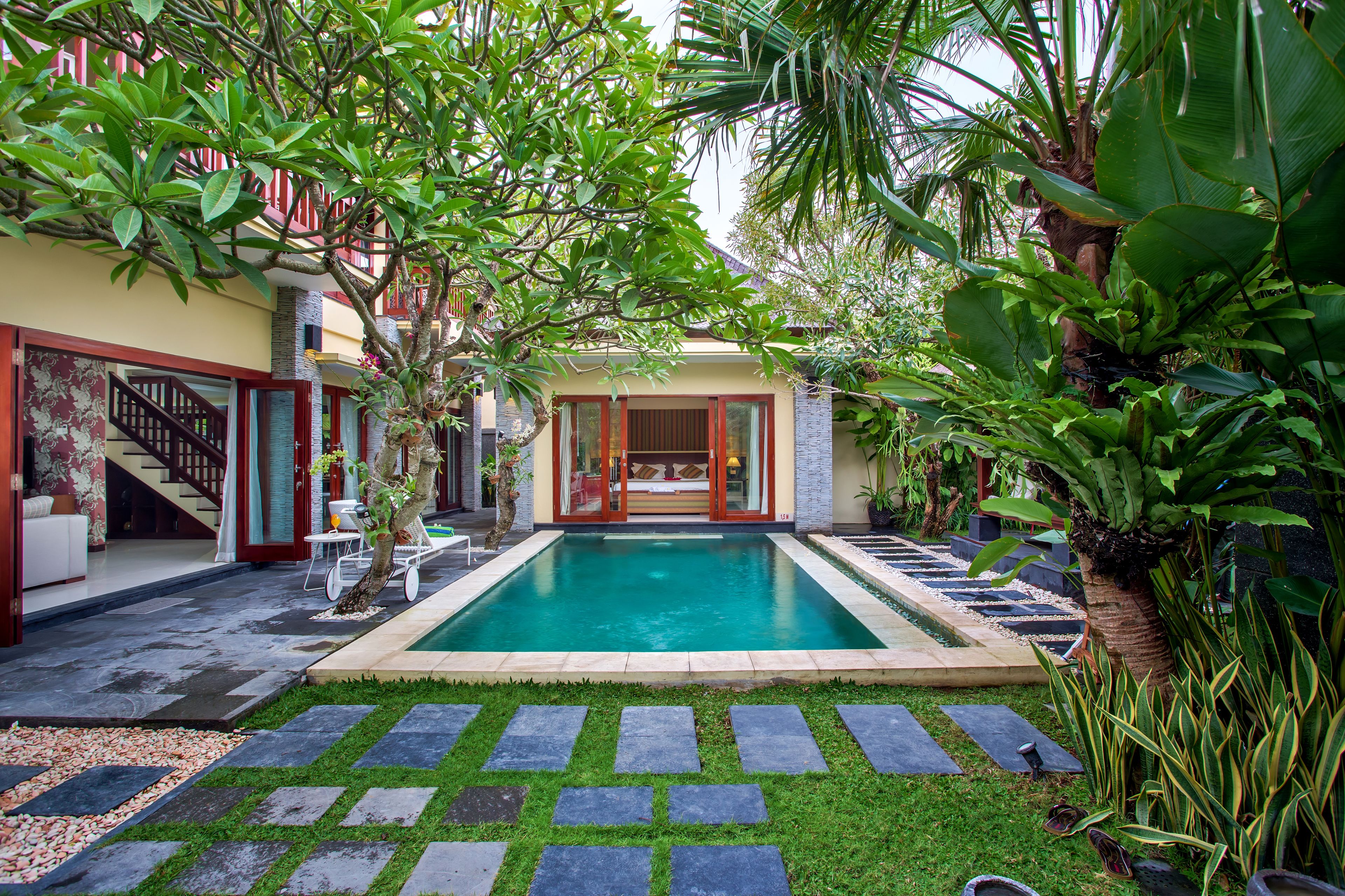 Una piscina rodeada por un jardín, con un patio y una escalera que lleva a un edificio.