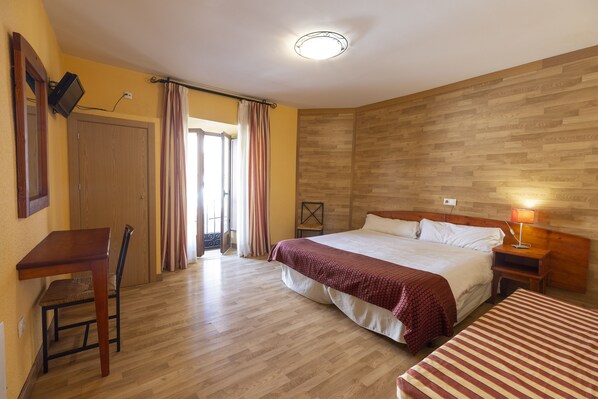 A hotel room with a wooden floor, a bed with a red and white bedspread, a desk with a chair, a nightstand with a lamp, and a wooden headboard.
