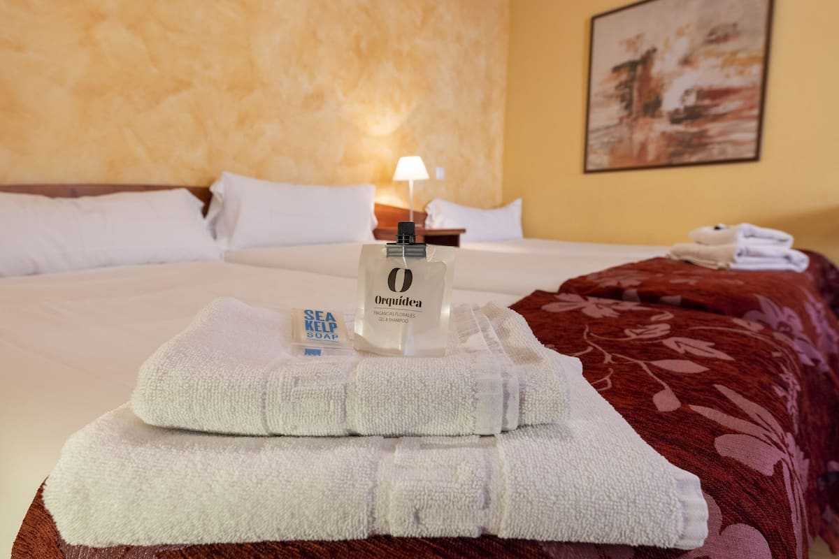 Un lit bien fait, avec du linge de lit blanc, une bouteille de gel douche Orquidea et une serviette blanche pliée posée sur le lit.