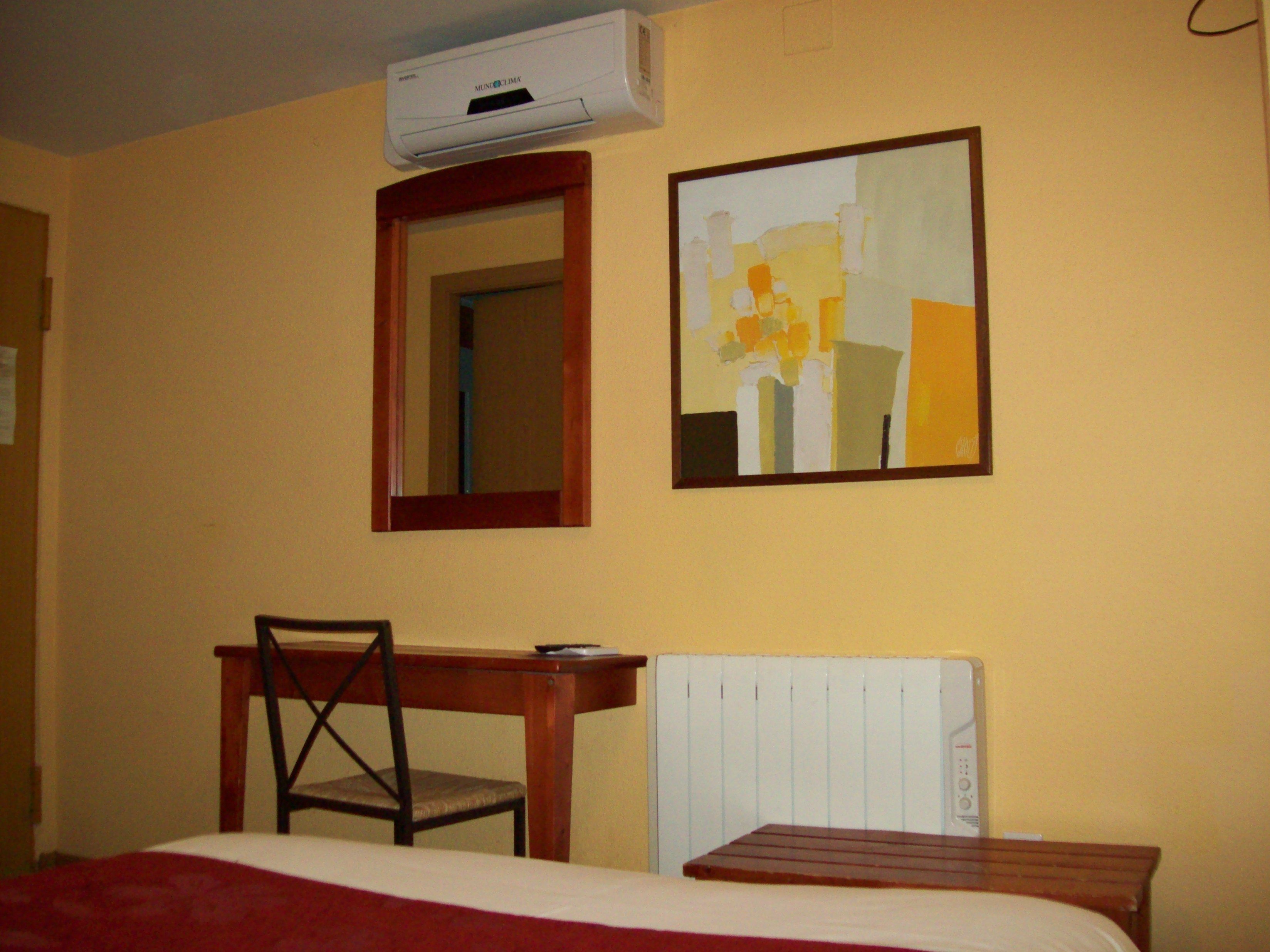 Habitación de hotel con cama, una silla de madera, escritorio, aire acondicionado, espejo y un cuadro en la pared.