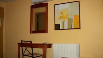 Habitación de hotel con cama, una silla de madera, escritorio, aire acondicionado, espejo y un cuadro en la pared.
