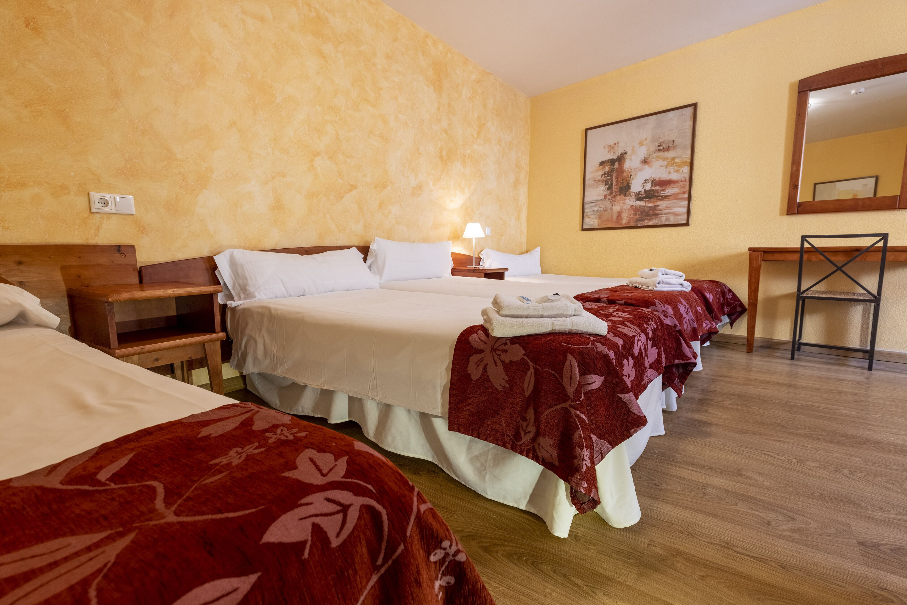 Habitación de hotel con tres camas, cada una con colcha floral roja, una mesita de noche de madera, una mesilla auxiliar y un cuadro colgado en la pared.