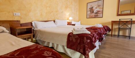Habitación de hotel con tres camas, cada una con colcha floral roja, una mesita de noche de madera, una mesilla auxiliar y un cuadro colgado en la pared.
