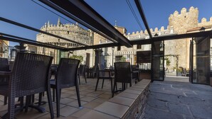 Een terras buiten met rieten stoelen en tafels, stenen vloeren en een uitzicht op een historisch stenen gebouw met torens.