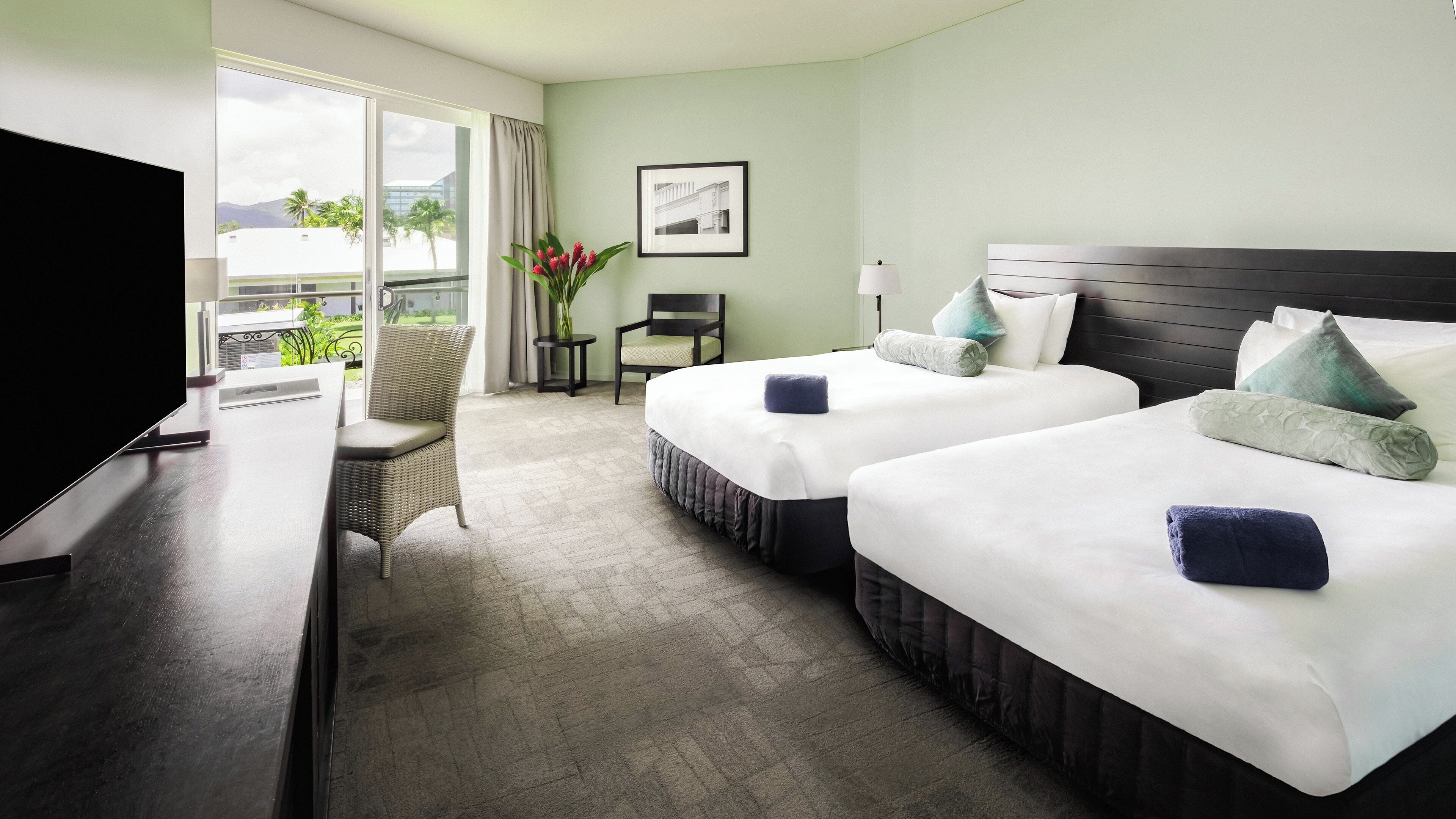 A hotel room with two beds, a TV, a chair, a small table, and a window with a view of palm trees.