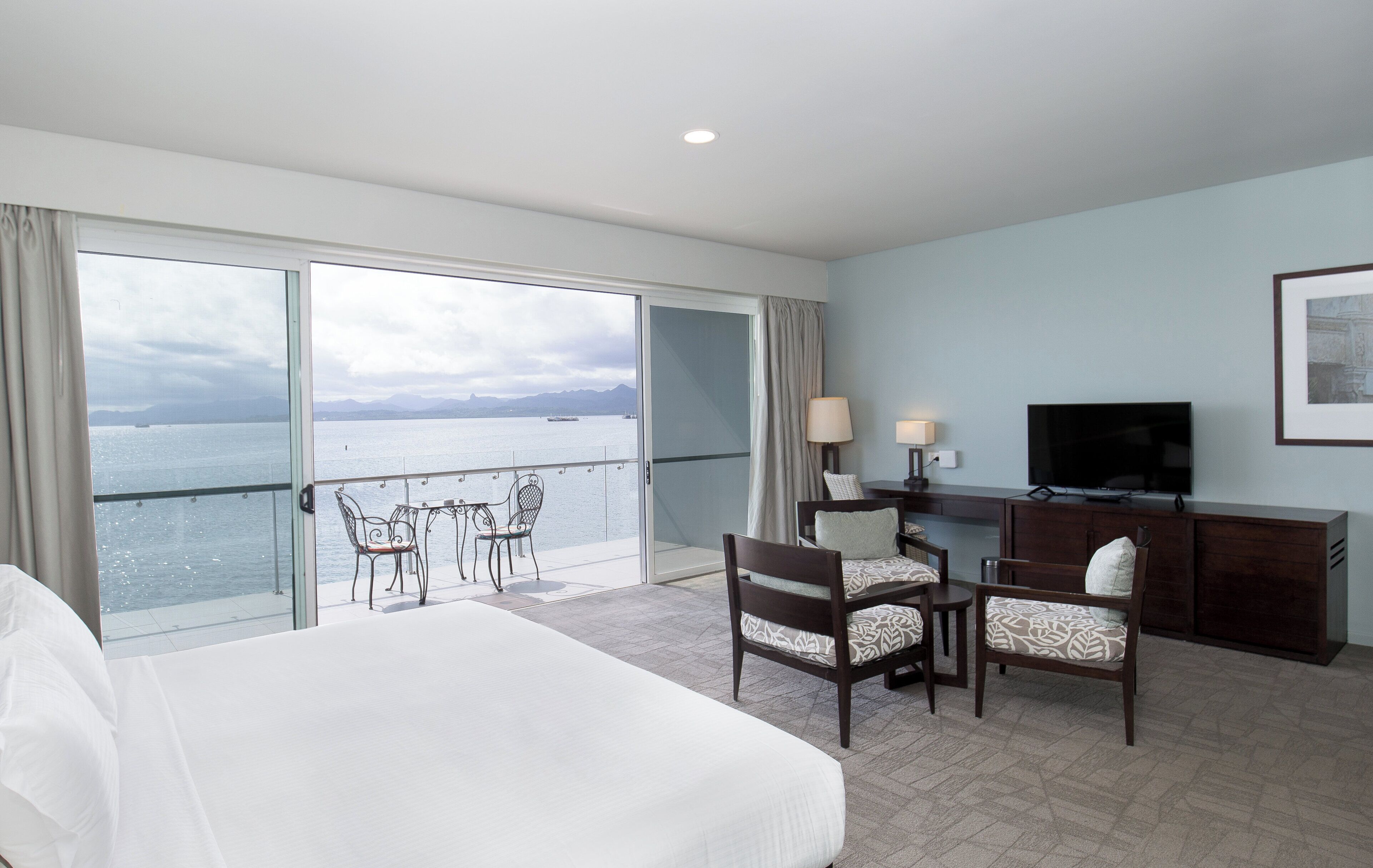 A hotel room with a large bed, a desk with a TV, a chair, and a view of the sea.