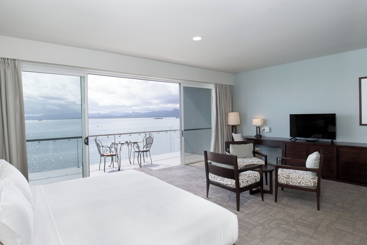 A hotel room with a large bed, a desk with a TV, a chair, and a view of the sea.