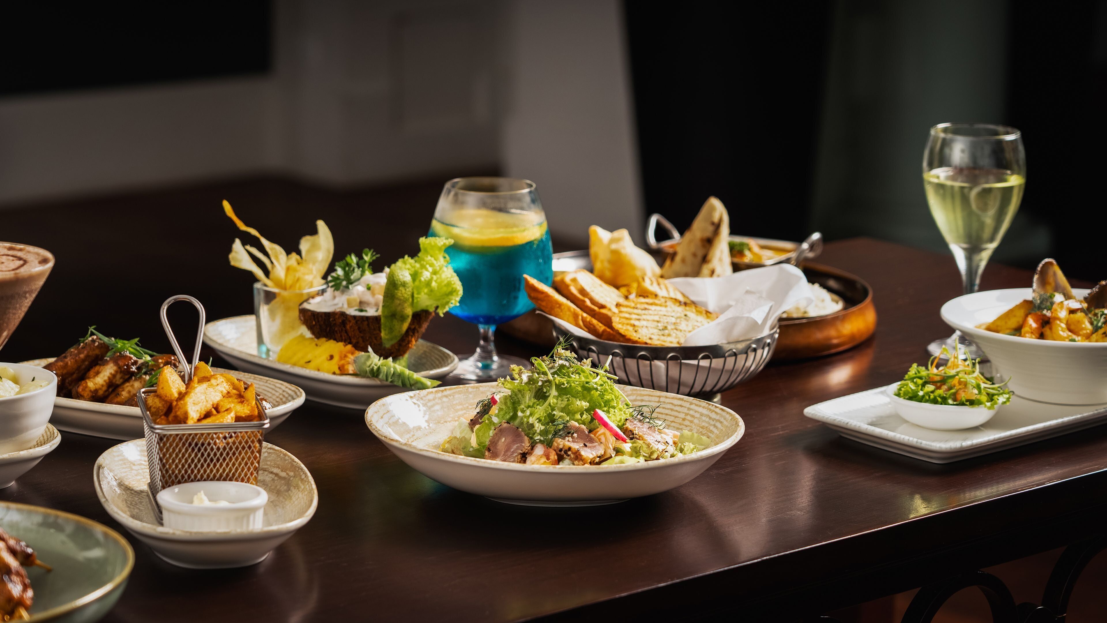A variety of dishes and drinks on a table, including a blue cocktail, bread, and a bowl of salad.