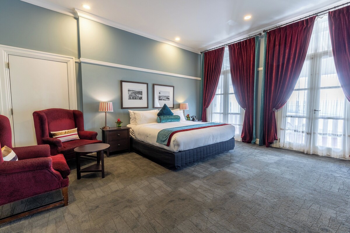 A hotel room with a bed, a red armchair, a small table, and two bedside lamps.