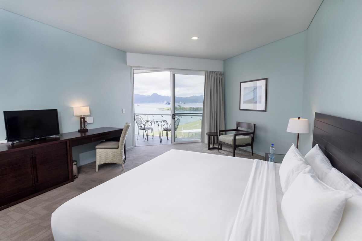 A hotel room with a large bed, a desk with a TV, a chair, a balcony with a view of the water and mountains.