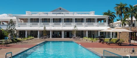A large pool area with a two-story building in the background, surrounded by palm trees and outdoor seating.