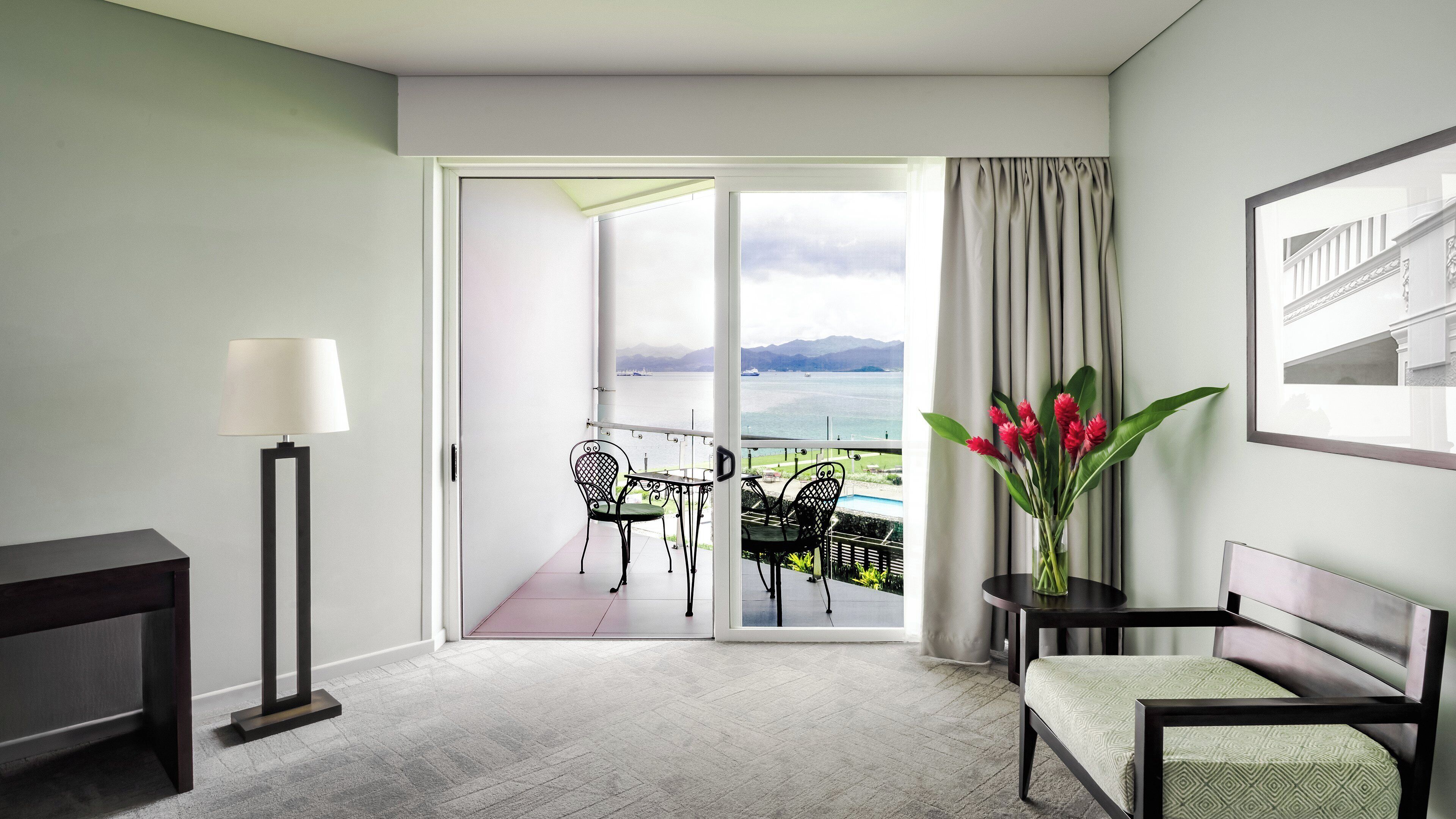 A hotel room with a balcony, a desk, a chair, a lamp, and a vase with flowers.