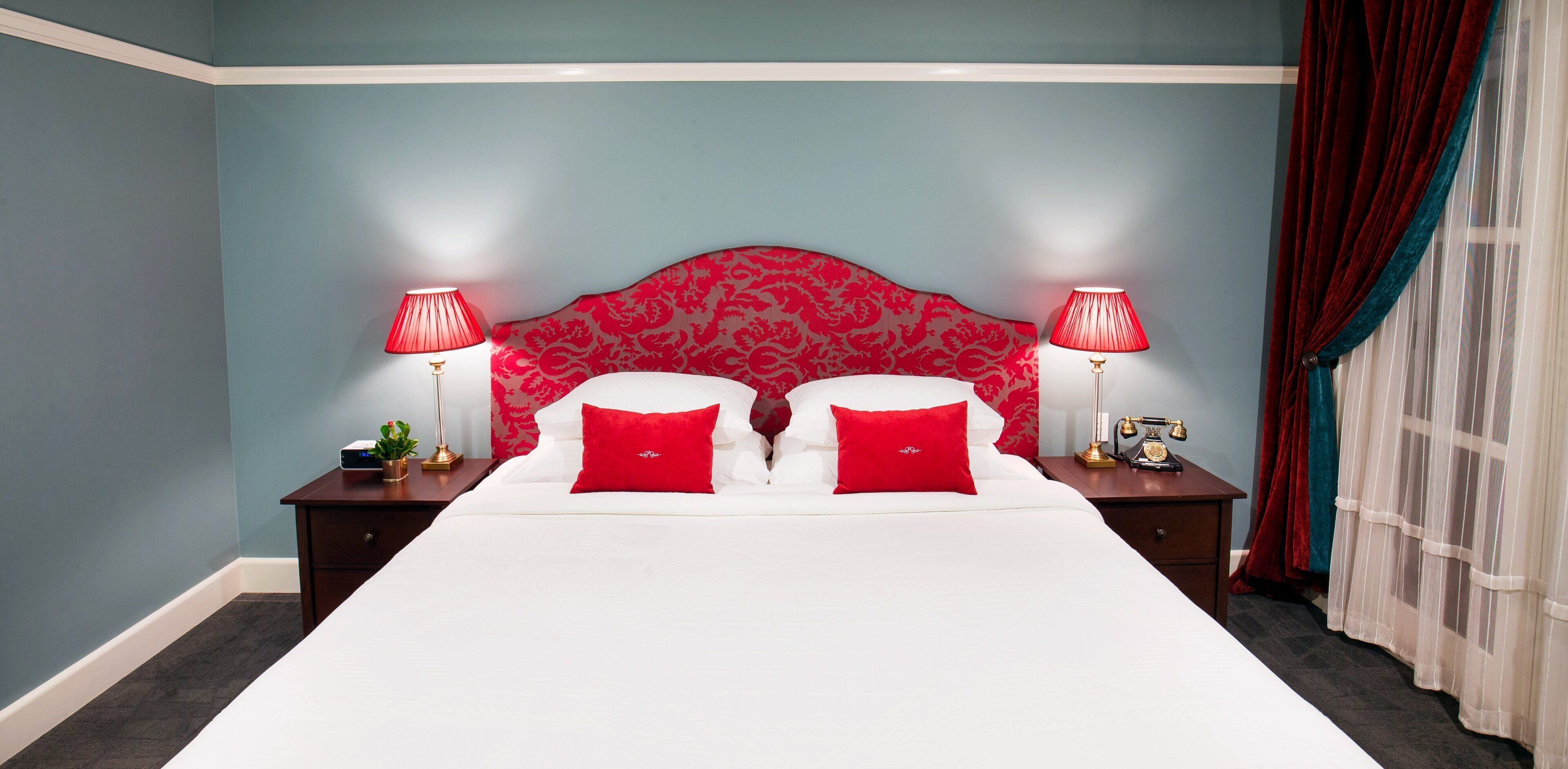 A hotel room with a large bed, two bedside tables, red lamps, and a red patterned headboard.