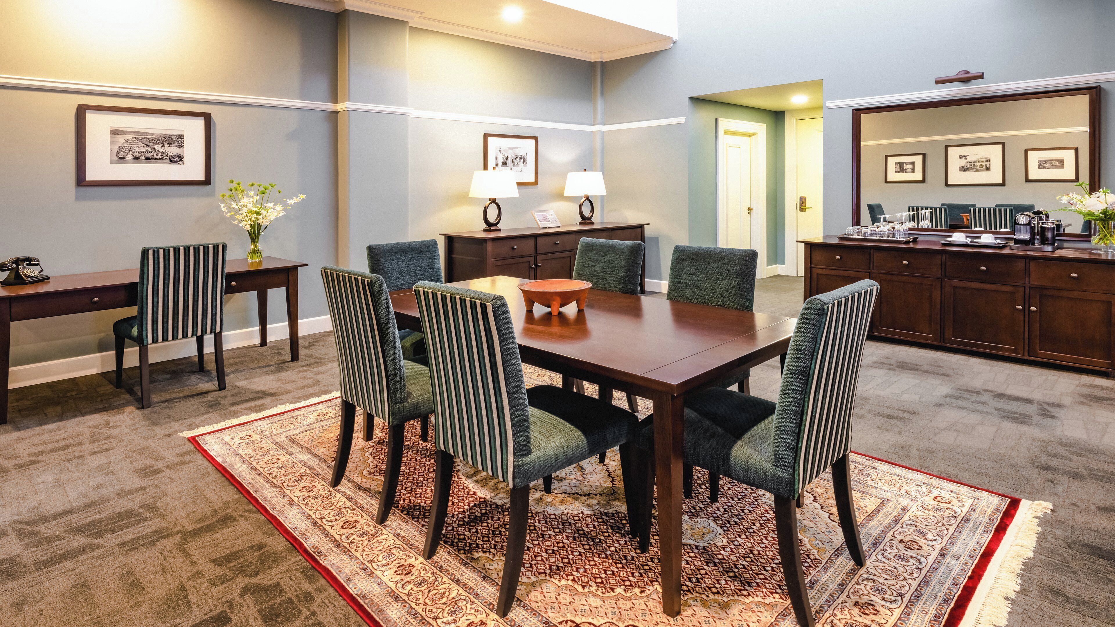 Uma sala de conferências com uma mesa de madeira, seis cadeiras, um tapete com padrão em vermelho e bege, uma pequena mesa lateral com uma luminária e quadros nas paredes.