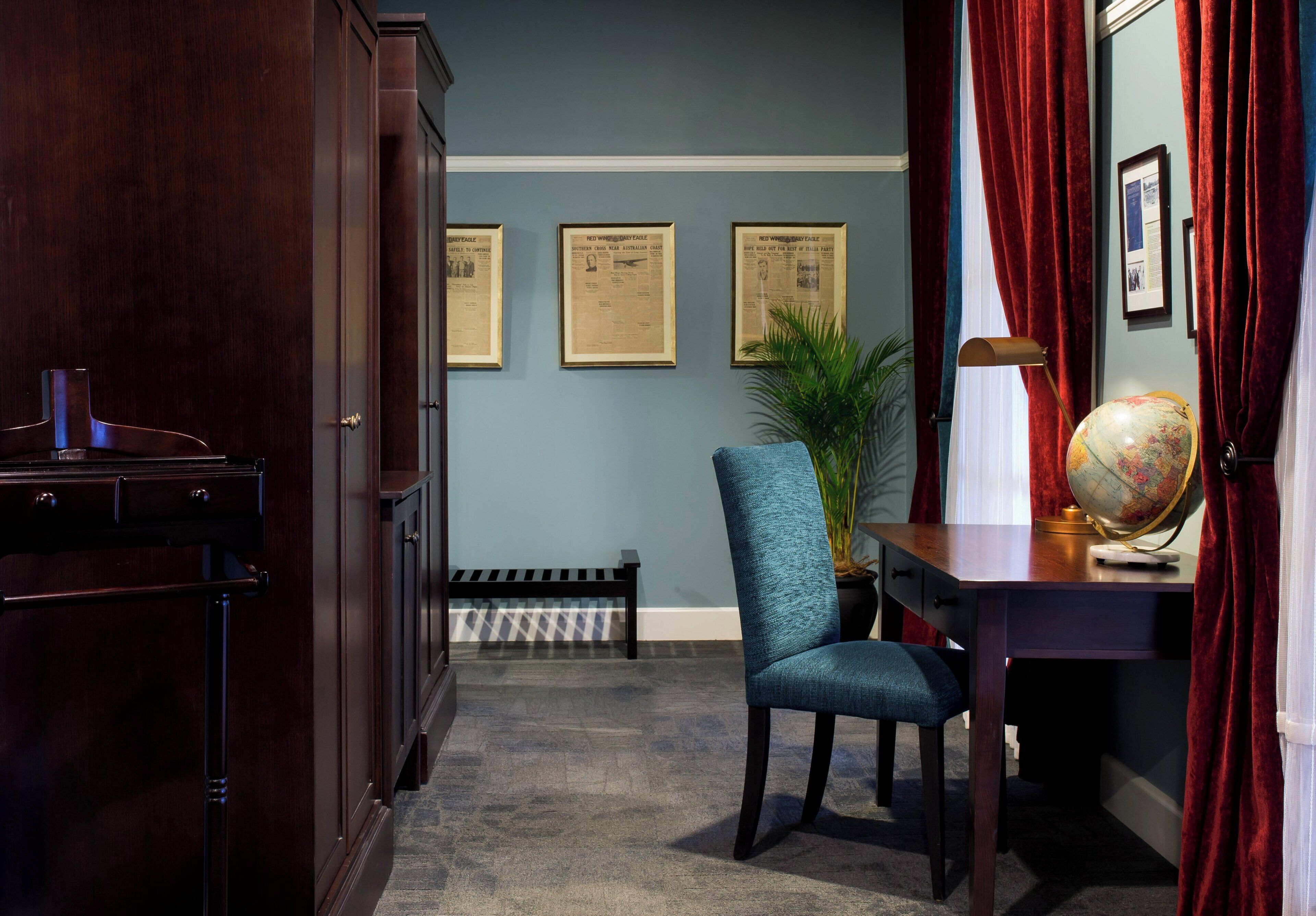 A room with a dark wood desk, a globe, a lamp, and a chair. There are framed certificates on the wall and a radiator below.