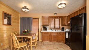 Una cocina con armarios de madera, un refrigerador negro, una mesa de comedor con sillas y una ventana con persianas.
