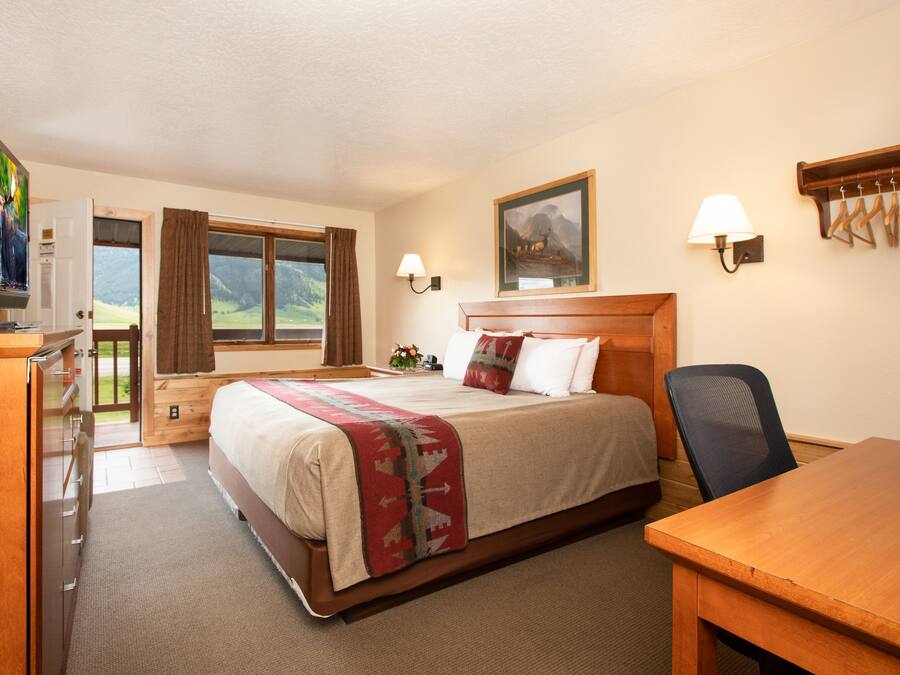 Quarto de hotel com cama, escrivaninha, televisão, janela com cortinas, um quadro na parede e um cabideiro.