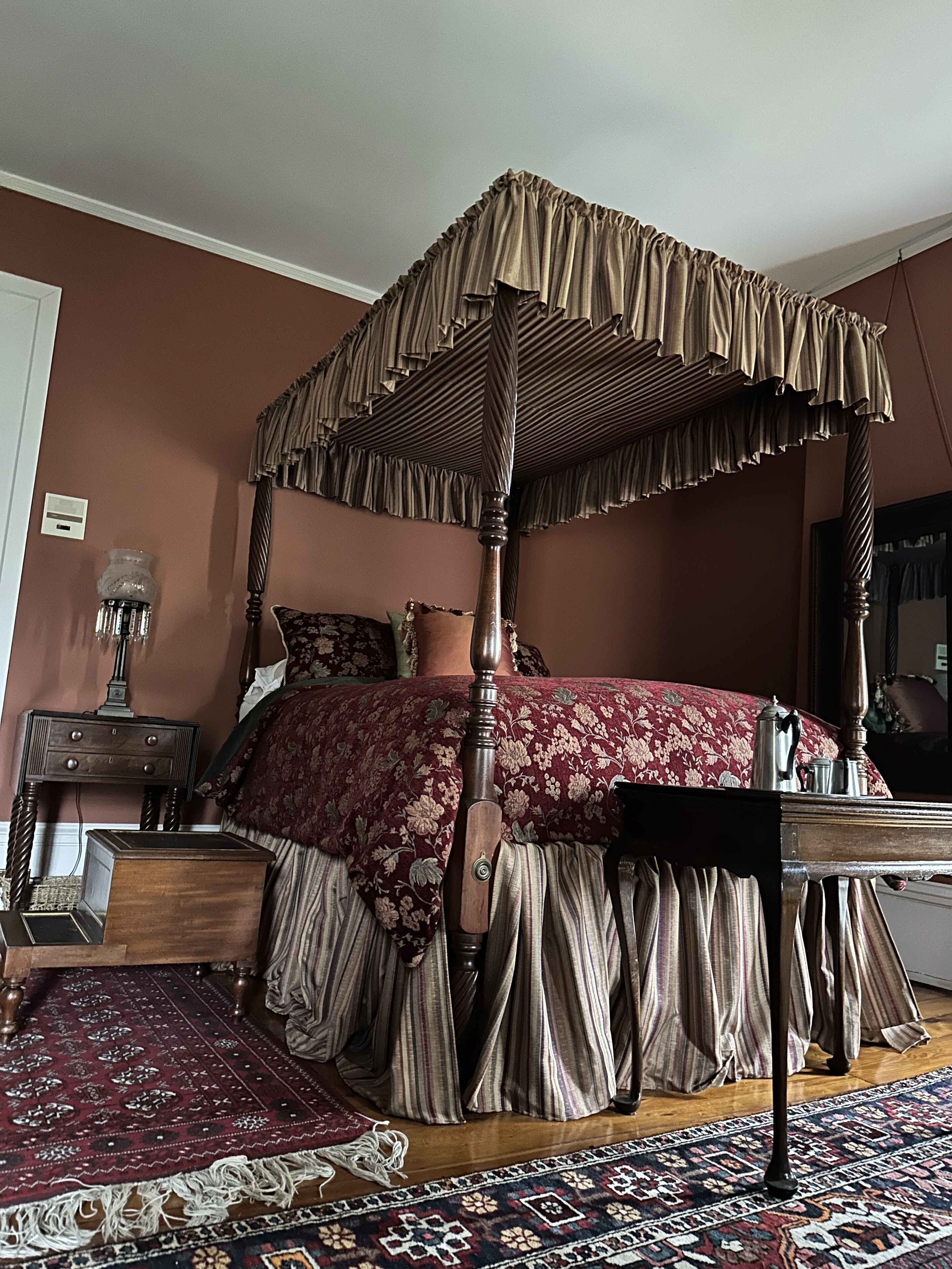 A four-poster canopy bed with a floral bedspread, a wooden nightstand with a lamp, and a patterned rug.