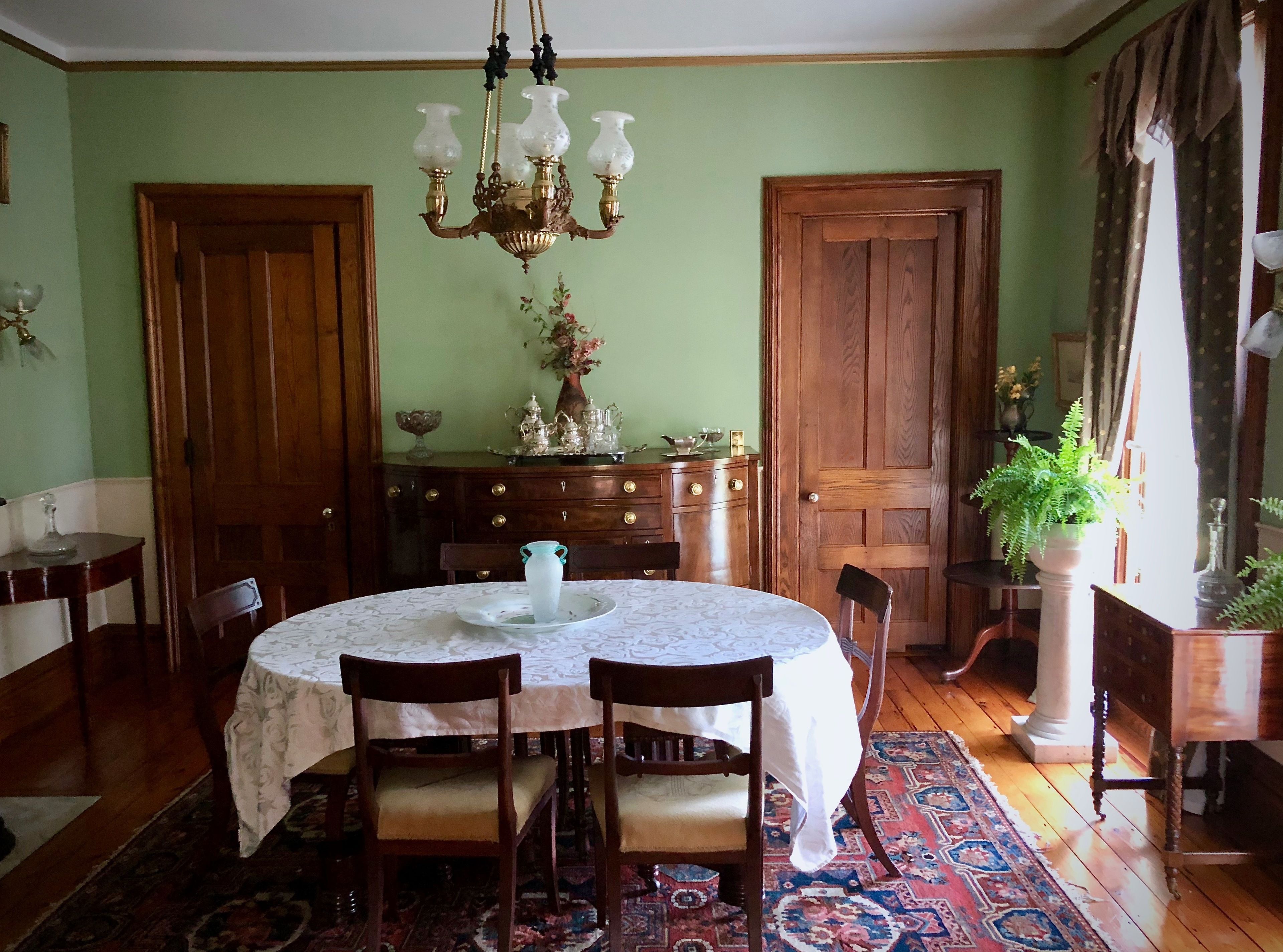 Une salle à manger avec une table ronde, des chaises, un lustre, des portes en bois, un meuble contenant des objets décoratifs et un tapis à motifs.