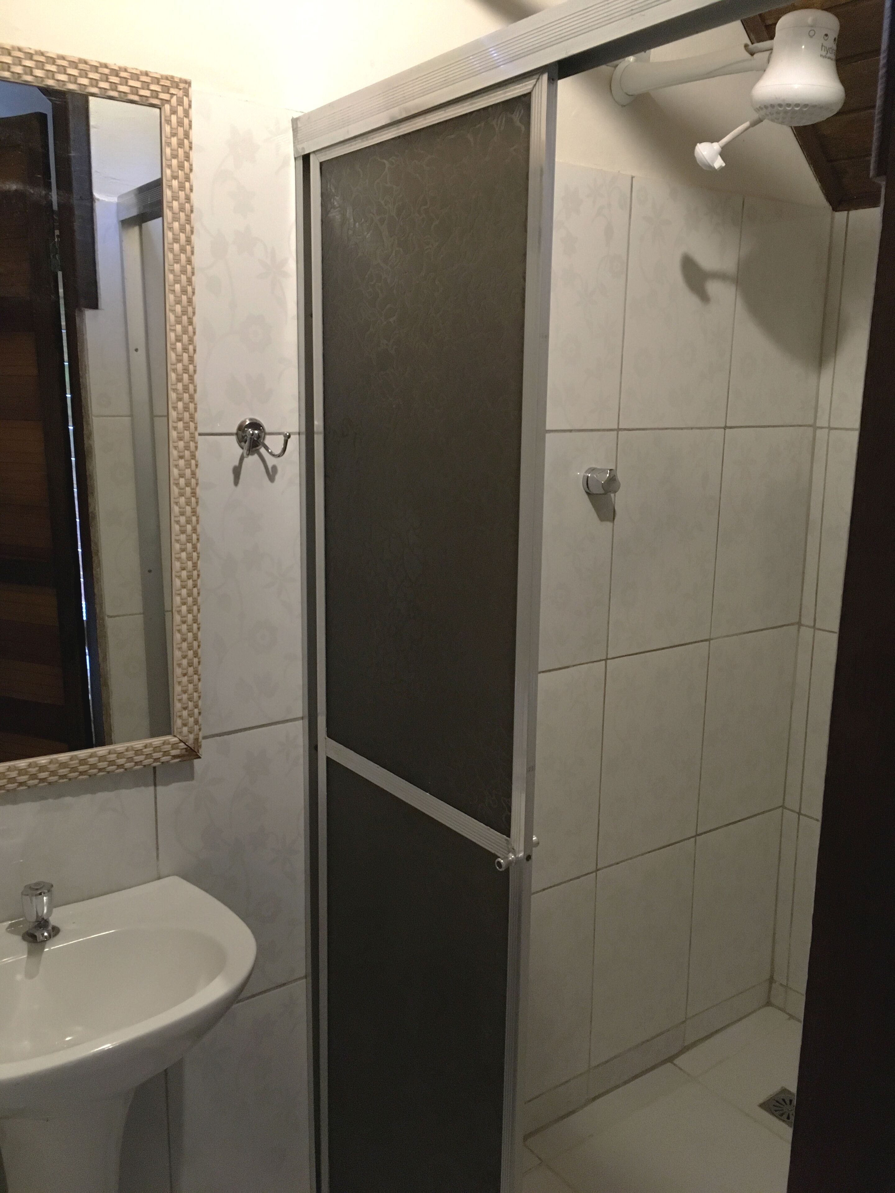 Ein Badezimmer mit Waschbecken in Weiß, einem Spiegel mit dekorativem Rahmen, einer Dusche mit Glastür und einer Deckenleuchte.